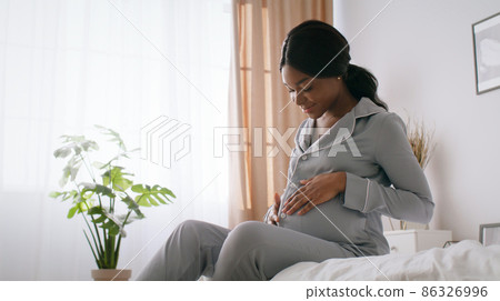 Young happy african american in pajamas sitting on bed and embracing belly, enjoying pregnancy time at home, free space Young happy african american in pajamas sitting on bed and embracing belly, enjoying pregnancy time at home, free space 86326996