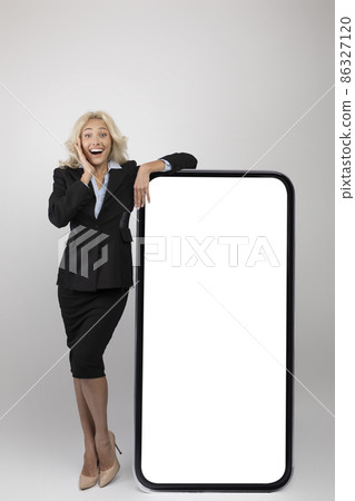Business app. Excited female entrepreneur in formal wear leaning at big blank smartphone with white screen, mockup Business app. Excited female entrepreneur in formal wear leaning at big blank smartphone with white screen, mockup 86327120