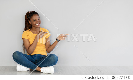 Cheery young black woman sitting cross legged on floor, pointing aside at free space against grey wall, banner 86327128