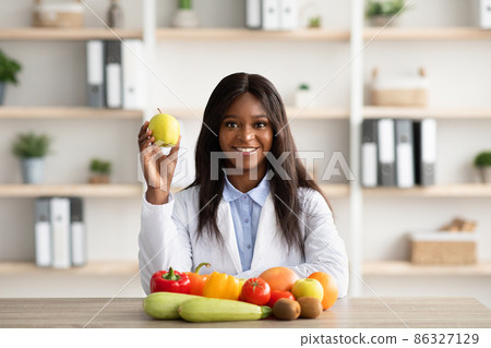Portrait of black female dietologist holding apple, recommending fresh fruits and vegetables, sitting in medical office Portrait of black female dietologist holding apple, recommending fresh fruits and vegetables, sitting in medical office 86327129