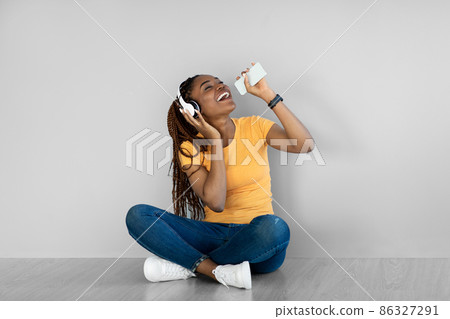 Cheerful black woman in headphones enjoying song in mobile audio app, listening to music, using smartphone as mic 86327291