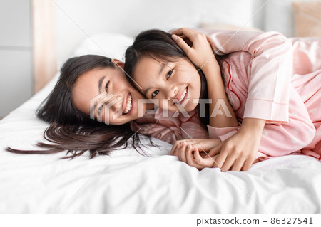 Glad happy pretty cute japanese young woman hug teen girl in pink pajama lie on bed and have fun in bedroom Glad happy pretty cute japanese young woman hug teen girl in pink pajama lie on bed and have fun in bedroom 86327541