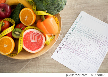 Weekly eating meal planner and plate of fresh fruits and vegetables nearby on table 86328065