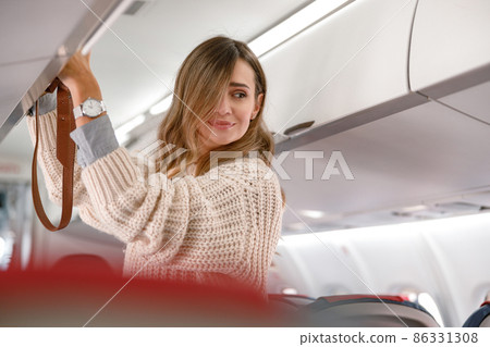 Young woman putting travel bag in overhead luggage bin Young woman putting travel bag in overhead luggage bin 86331308