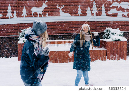 Two young women play snowballs and laugh while walking through a snowy town Two young women play snowballs and laugh while walking through a snowy town 86332824