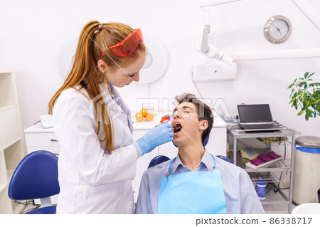 Ginger woman in white robe and latex gloves using mirror to examine teeth of man while working in modern office of dental clinic 86338717