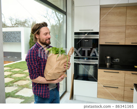 Charming young man enters the kitchen holding paper craft eco bag with food. Handsome guy with cardboard sack full of healthy food and whole grain bread, stands by kitchen countertop in spacious villa Charming young man enters the kitchen holding paper craft eco bag with food. Handsome guy with cardboard sack full of healthy food and whole grain bread, stands by kitchen countertop in spacious villa 86341165