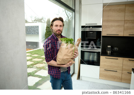 Handsome Caucasian man, charming guy, stands in a home kitchen with a cardboard bag full of healthy food and whole grain bread, looks at camera against the background of windows overlooking the garden 86341166