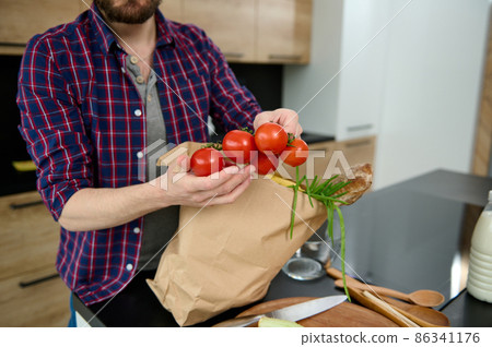 Close-up of a Caucasian man taking out vegetables and healthy food from a paper eco bag, holding a sprig of fresh tomatoes in his hands, putting it on the kitchen table. Healthy eating concept 86341176