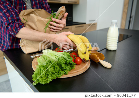 Overhead view of an unrecognizable man unpacking eco cardboard bag with healthy raw vegan food and a whole grain baguette, putting it on a kitchen table. Healthy food and lifestyles concept 86341177