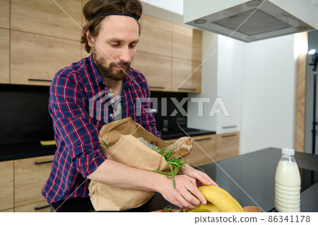 Handsome Caucasian man unpacking eco cardboard bag with healthy raw vegan food and a whole grain baguette, bananas and putting ingredients on a kitchen table. Healthy food and lifestyles concept 86341178