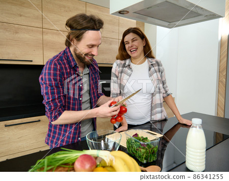 Happy heterosexual Caucasian couple of a handsome man and beautiful delightful pregnant woman standing together by kitchen countertop in their spacious villa and and cooking healthy fresh lunch 86341255