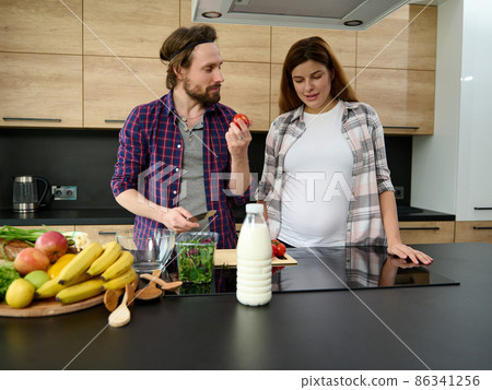 Caucasian young heterosexual couple in love, delightful pregnant woman standing next to her husband who showing her a fresh tomato while chopping vegetables for salad, cooking together at home kitchen 86341256