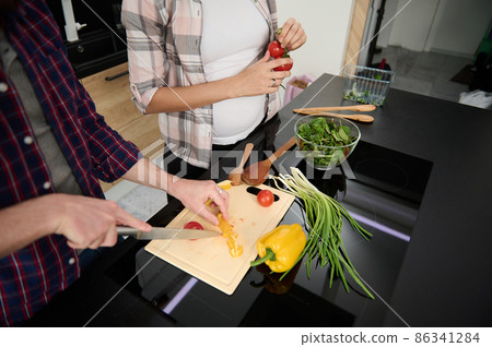 Close-up of hands chopping vegetables on a cutting board and cropped view to a pregnant woman holding sprig with fresh tomatoes while enjoying cooking together with her husband at home kitchen Close-up of hands chopping vegetables on a cutting board and cropped view to a pregnant woman holding sprig with fresh tomatoes while enjoying cooking together with her husband at home kitchen 86341284