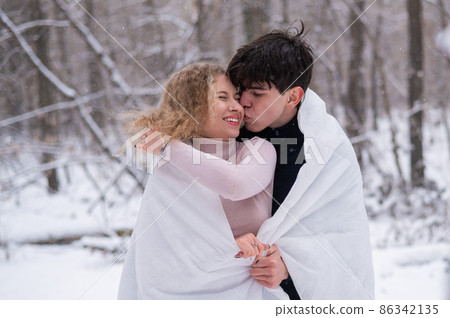 A young couple walks in the park in winter. The guy and the girl are kissing wrapped in a white blanket outdoors. A young couple walks in the park in winter. The guy and the girl are kissing wrapped in a white blanket outdoors. 86342135