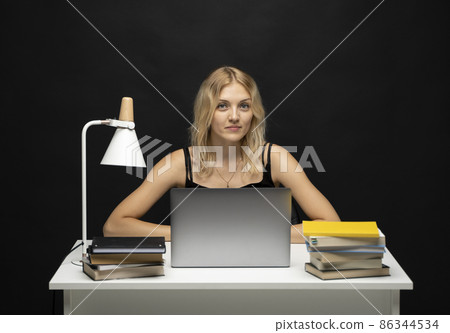 Smiling business woman working with a laptop isolated on a black background. Portrait of a pretty young woman studying while sitting at the table with grey laptop computer, notebook. 86344534