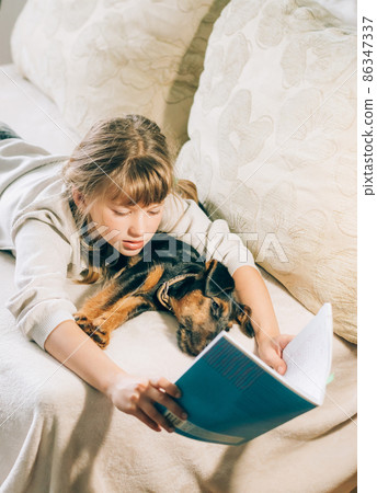 A girl and small dog puppy lie on the couch engaged in reading training. The concept of friendship between man and dog 86347337