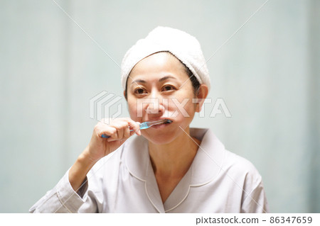 A middle-aged woman in pajamas who wraps a towel around her head and brushes her teeth 86347659