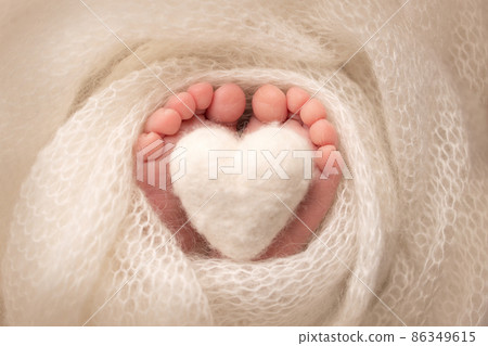 The tiny foot of a newborn baby. Soft feet of a new born in a white wool blanket. Close up of toes, heels and feet of a newborn. Knitted white heart in the legs of a baby. Macro photography.  86349615