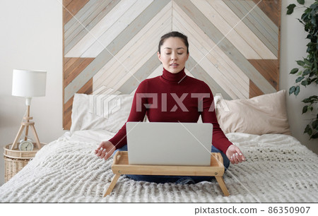 Relaxed mixed race young asian woman sitting on her bed with folded in mudra gesture hands, reducing stress during workday, chilling or doing yoga breathing exercises, working from home with laptop 86350907