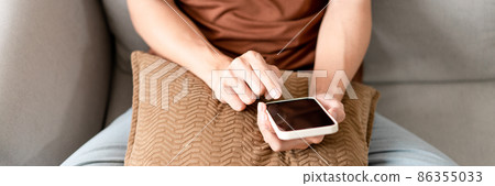 Technology Concept The man sitting next to a pot of plant, putting a small pillow on his lap, and checking something on his smartphone Technology Concept The man sitting next to a pot of plant, putting a small pillow on his lap, and checking something on his smartphone 86355033
