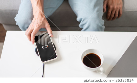Technology Concept A person with his light blue jeans sitting on the couch and trying to charge his smartphone on the wireless battery charger 86355034