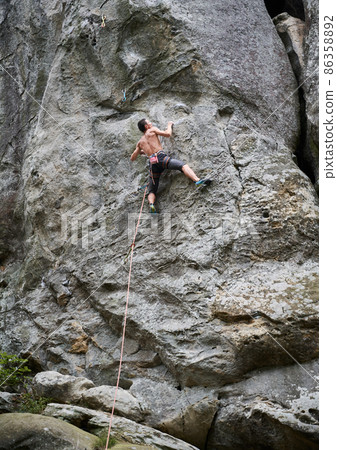 Young man climber climbing difficult route on a high boulder with rope. Fearless climber training rock climbing on summer day. Concept of extreme sport, adventures and active lifestyle. 86358892