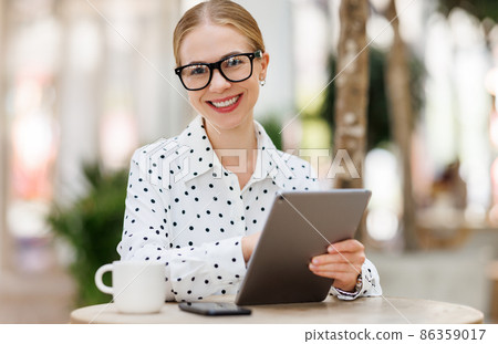 Lovely student girl in glasses sitting with tablet and cup of takeaway coffee outside on terrace Lovely student girl in glasses sitting with tablet and cup of takeaway coffee outside on terrace 86359017