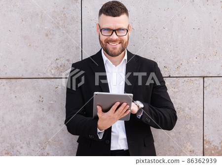 Cheerful businessman in suit using digital tablet while standing outside against concrete wall 86362939