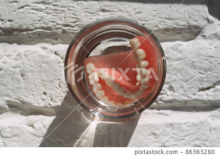 Prosthesis in a glass with a solution. Dental prosthesis care. Full removable plastic denture of the jaws. Acrylic denture. Lower jaw with false teeth. Dentures or false teeth, close-up. Prosthesis in a glass with a solution. Dental prosthesis care. Full removable plastic denture of the jaws. Acrylic denture. Lower jaw with false teeth. Dentures or false teeth, close-up. 86363280