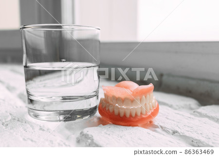 A denture in a glass of water. Dental prosthesis care. Full removable plastic denture of the jaws. Two acrylic dentures. Upper and lower jaws with fake teeth. Dentures or false teeth, close-up. A denture in a glass of water. Dental prosthesis care. Full removable plastic denture of the jaws. Two acrylic dentures. Upper and lower jaws with fake teeth. Dentures or false teeth, close-up. 86363469