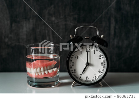 A denture in a glass of liquid standing on the bedside table in the early morning. Prosthesis in a glass with a solution. Dental prosthesis care. Dentures or false teeth. Copy space A denture in a glass of liquid standing on the bedside table in the early morning. Prosthesis in a glass with a solution. Dental prosthesis care. Dentures or false teeth. Copy space 86363735
