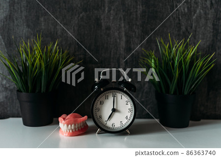 Full removable plastic prosthetic jaw and alarm clock. A denture next to the clock on the bedside table in the early morning. Dentures or false teeth on the bedside table. Copy space Full removable plastic prosthetic jaw and alarm clock. A denture next to the clock on the bedside table in the early morning. Dentures or false teeth on the bedside table. Copy space 86363740