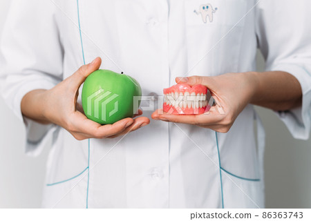A false denture and a green apple in the hands of a dentist. Dental prosthesis care. A denture and an apple in the hands of an orthopedic dentist. Dental care. Prosthetics. False teeth A false denture and a green apple in the hands of a dentist. Dental prosthesis care. A denture and an apple in the hands of an orthopedic dentist. Dental care. Prosthetics. False teeth 86363743