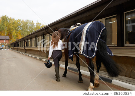 Female horseman going with her brown horse outdoor. Concept of animal care. Rural rest and leisure. Idea of green tourism. Young european woman wearing helmet and uniform 86363824