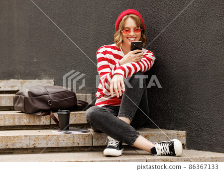 Smiling student girl using mobile phone while sitting outside with takeaway coffee before in morning 86367133