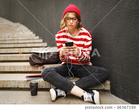 Moody upset student girl in earphones sitting outside and texting friends on cell phone 86367199