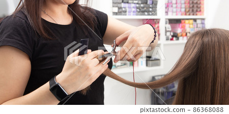 Professional hair stylist holding comb and hot thermal scissors cutting tips of long straight hair lock closeup. Professional hair stylist holding comb and hot thermal scissors cutting tips of long straight hair lock closeup. 86368088