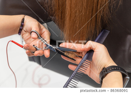 Professional hair stylist holding comb and hot thermal scissors cutting tips of long straight hair lock closeup. 86368091