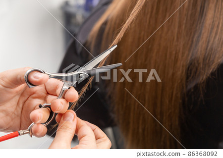Professional hair stylist holding comb and hot thermal scissors cutting tips of long straight hair lock closeup. Professional hair stylist holding comb and hot thermal scissors cutting tips of long straight hair lock closeup. 86368092