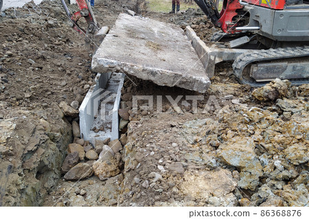 The excavator installs concrete trays for drainage of rainwater in rural areas, concrete products for drainage of water. The excavator installs concrete trays for drainage of rainwater in rural areas, concrete products for drainage of water. 86368876