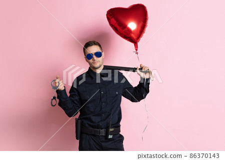 Young charming policeman officer with heart shape balloon wearing sunglasses isolated on pink background. St, Valentine's day Young charming policeman officer with heart shape balloon wearing sunglasses isolated on pink background. St, Valentine's day 86370143