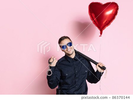 Young charming policeman officer with heart shape balloon wearing sunglasses isolated on pink background. St, Valentine's day Young charming policeman officer with heart shape balloon wearing sunglasses isolated on pink background. St, Valentine's day 86370144