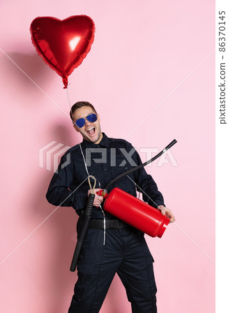 Comic portrait of young charming policeman officer having fun with heart shape balloon and fire extinguisher isolated on pink background. St, Valentine's day Comic portrait of young charming policeman officer having fun with heart shape balloon and fire extinguisher isolated on pink background. St, Valentine's day 86370145