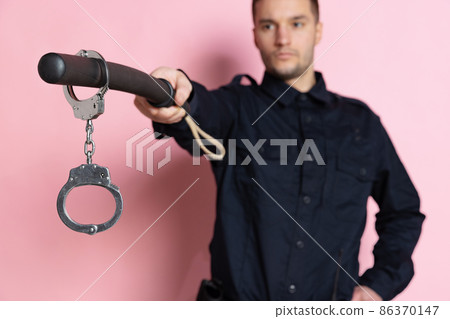 One handsome policeman officer wearing black uniform with club and handcuffs isolated on pink background. One handsome policeman officer wearing black uniform with club and handcuffs isolated on pink background. 86370147