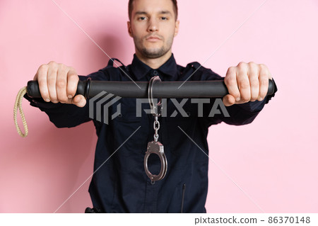 One handsome policeman officer wearing black uniform with club and handcuffs isolated on pink background. One handsome policeman officer wearing black uniform with club and handcuffs isolated on pink background. 86370148