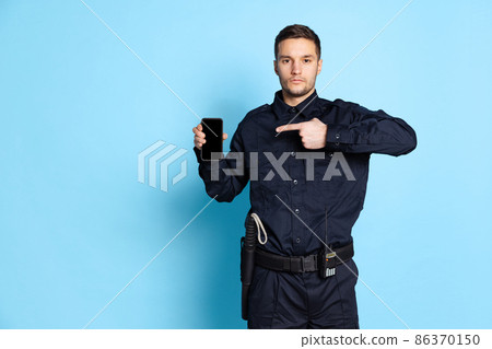 Portrait of young man, policeman officer wearing black uniform using phone isolated on blue background. Concept of job, caree, law and order. Portrait of young man, policeman officer wearing black uniform using phone isolated on blue background. Concept of job, caree, law and order. 86370150