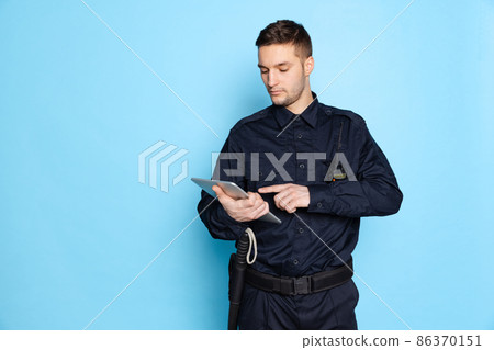 Portrait of young man, policeman officer wearing black uniform using tablet isolated on blue background. Concept of job, caree, law and order. Portrait of young man, policeman officer wearing black uniform using tablet isolated on blue background. Concept of job, caree, law and order. 86370151