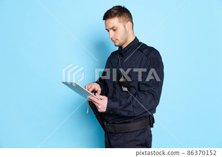 Portrait of young man, policeman officer wearing black uniform using tablet isolated on blue background. Concept of job, caree, law and order. Portrait of young man, policeman officer wearing black uniform using tablet isolated on blue background. Concept of job, caree, law and order. 86370152