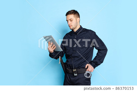 Portrait of young man, policeman officer wearing black uniform using tablet isolated on blue background. Concept of job, caree, law and order. Portrait of young man, policeman officer wearing black uniform using tablet isolated on blue background. Concept of job, caree, law and order. 86370153